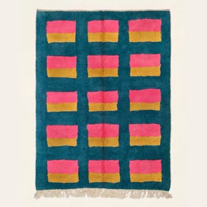Puede incluir: Una alfombra rectangular con fondo turquesa y un patrón repetitivo de rectángulos rosas y dorados. La alfombra tiene flecos en el borde inferior. El diseño es moderno y geométrico, ideal para el hogar.