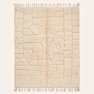 Puede incluir: Alfombra rectangular color crema con un patrón geométrico. La alfombra presenta un diseño texturizado con varias formas y líneas, y tiene flecos en los bordes superior e inferior. Adecuada para una sala de estar o dormitorio.