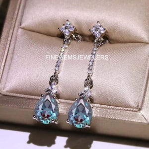 May include: A pair of silver drop earrings. Each earring has a teardrop-shaped, teal gemstone, with smaller clear stones and a diamond-shaped top. The text "FINEGEMSJEWELERS" is visible.