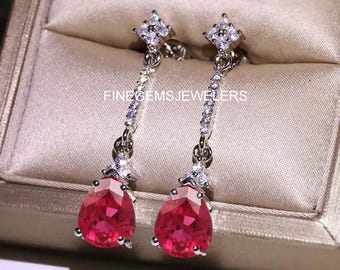 May include: Silver drop earrings with a vibrant red, pear-shaped gemstone. The earrings have a delicate chain of small, clear stones and a square top with clear stones. The text "FINEGEMSJEWELERS" is visible. The earrings are presented in a beige box.