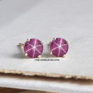 May include: A pair of gold stud earrings featuring a star ruby gemstone in each earring. The ruby is a deep pink colour with a star-like pattern.