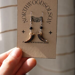 May include: Bear claw earrings with silver hooks and black bead detailing, displayed on a brown card. The card features the text "NORTHWOODSOLSON" and a butterfly design. The earrings are a unique accessory.