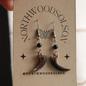 May include: Dangle earrings with natural claw pendants, silver-tone metal findings, and black beads. The earrings are presented on a brown card with the text "NORTHWOODSOLSON" and a butterfly illustration.