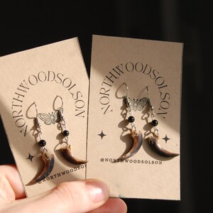 May include: Two pairs of earrings with a claw design. Each earring features a curved, brown claw pendant, black beads, and silver-coloured hooks. The earrings are displayed on tan cards with the text "NORTHWOODSOLSON" and a butterfly graphic.