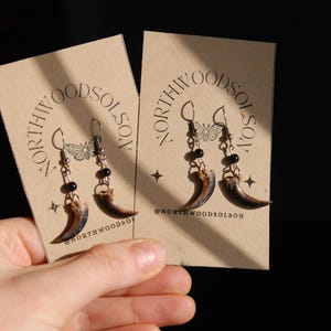 May include: Earrings with claw pendants, black beads, and silver hooks, presented on a tan card. The card displays the text "NORTHWOODSOLSON" and a butterfly design. The brand's Instagram handle is also shown on the card.