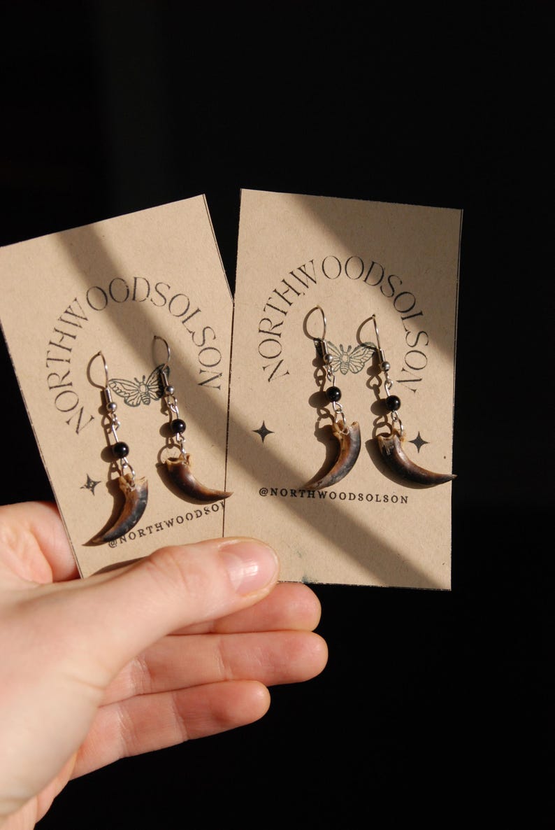 May include: Earrings with claw pendants, black beads, and silver hooks, presented on a tan card. The card displays the text "NORTHWOODSOLSON" and a butterfly design. The brand's Instagram handle is also shown on the card.