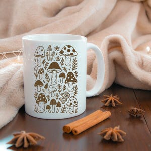 May include: White ceramic mug featuring a brown illustration of various mushrooms and foliage. The design includes different mushroom types with dotted and patterned caps. The mug has a curved handle and is set against a soft, neutral background.