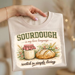 Könnte beinhalten: Beiges Rundhals-T-Shirt mit dem grünen Text "SOURDOUGH is my love language" und einer Illustration eines Sauerteigglases, eines Brotlaibs und Blumen. Der Satz "rooted in simple living" ist ebenfalls enthalten.