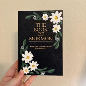 May include: A black book titled "THE BOOK OF MORMON" with the text "ANOTHER TESTAMENT OF JESUS CHRIST." The cover is decorated with white daisies with yellow centers and green leaves.