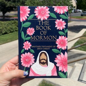 May include: A navy blue book titled "The Book of Mormon" with pink floral accents and the text "Another Testament of Jesus Christ." The cover features a depiction of Jesus. The book is held in front of a blurred outdoor background.