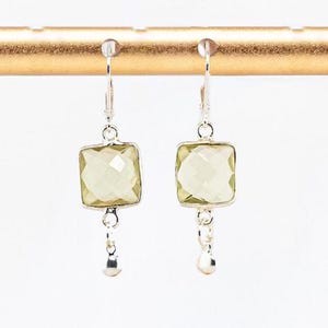 May include: A pair of silver leverback earrings featuring square, faceted, light green gemstones. Each earring has a small, round silver charm dangling from the bottom of the square stone, suspended from a silver wire hook mechanism.