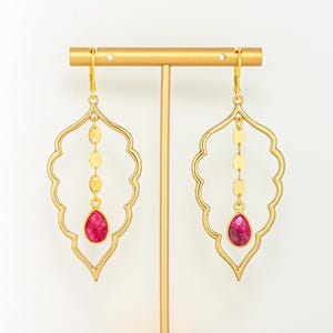 May include: A pair of gold-colored earrings with a leaf-shaped design. Each earring features a dangling chain of small gold circles and a teardrop-shaped, magenta-colored stone. The earrings are displayed on a gold stand against a white background.