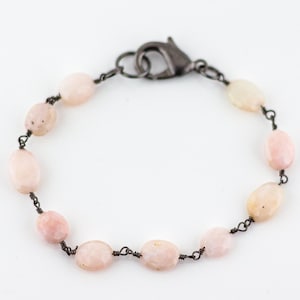 May include: A bracelet featuring a series of smooth, oval-shaped pale pink stones linked together with dark gunmetal wire. The piece is secured with a dark metal lobster claw clasp, creating a delicate and simple design with a neutral color palette.