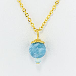 May include: A gold-colored chain necklace with a pendant featuring a large, round, light blue bead and a smaller, light blue bead below it. The beads have a marbled appearance. The pendant is attached to the chain with a decorative gold-colored bail.
