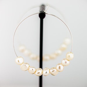 May include: A single silver hoop earring with a row of creamy white pearls. The pearls are evenly spaced along the bottom half of the hoop. The earring is displayed against a white background.