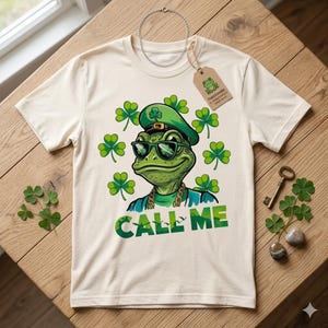 Pode incluir: Uma t-shirt de cor creme com o gráfico de um sapo verde a usar óculos de sol, uma corrente dourada e um chapéu verde com um trevo. O sapo está rodeado por trevos verdes e o texto "CALL ME" está impresso em letras grandes abaixo.