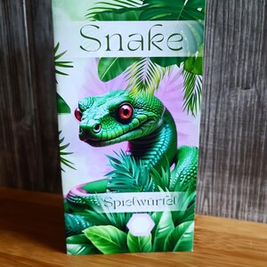 Könnte beinhalten: Eine rechteckige Schachtel mit einer lebendigen Illustration einer grünen Schlange mit roten Augen, umgeben von üppigem grünen Laub. Das Wort "Snake" steht oben, mit "Spielwürfel" unten. Der Hintergrund hat ein tropisches Thema.
