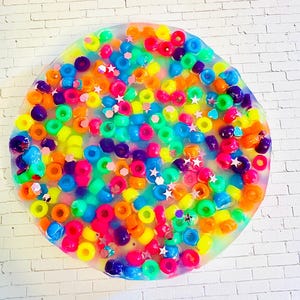 May include: A circular, clear resin coaster filled with colorful beads in shades of pink, orange, yellow, green, blue, and purple. Small star-shaped glitter pieces are scattered throughout the design. The coaster is set against a white brick wall.