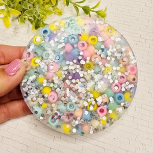 May include: A round, clear coaster filled with colorful beads in pastel shades of pink, yellow, blue, and green. Silver glitter is scattered throughout the coaster, adding sparkle. The coaster is held by a person.