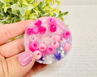 May include: A round, clear resin object filled with pink and purple beads. The beads vary in size and shape, with some appearing to be small cylinders and others round. The object is held in a hand.