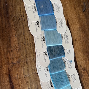 May include: A collection of blue silk thread samples, each labeled with color codes and measurements. The samples range in shades from deep to light blue, with the labels indicating the thread is 100% pure silk and spun in Japan.