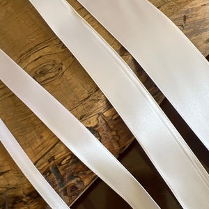 May include: Several white satin ribbons of varying widths are arranged diagonally across a wooden surface. The ribbons have a smooth, glossy texture and appear to be new. The ribbons are likely used for crafts, gift wrapping, or decorative purposes.