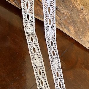 May include: Two white lace trims with a delicate floral pattern and oval cutouts. The trims are laid out on a wooden surface.