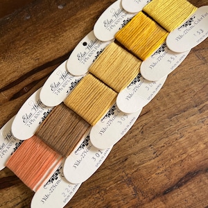 May include: A collection of Elsa Williams 7-Ply 100% Pure Silk embroidery floss in various shades of yellow, orange, and brown. Each spool is labeled with the color number and 3 yds. 275cm. of thread. The spools are arranged on a wooden surface.