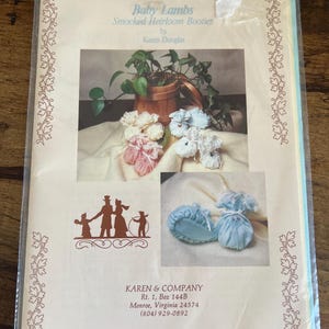 May include: A package featuring the text "Baby Lambs Smocked Heirloom Booties by Karen Douglas." The image shows examples of baby booties in pink, white, and blue, along with a family silhouette and a wooden planter with greenery.