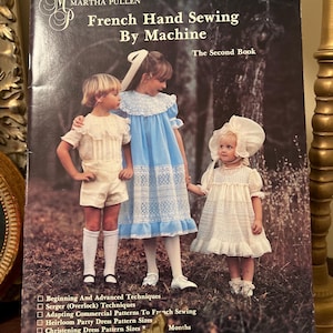Puede incluir: Una portada de libro vintage titulada "French Hand Sewing By Machine" de Martha Pullen. La portada presenta a tres niños con vestidos y pantalones cortos vintage. El subtítulo del libro es "The Second Book". La portada también enumera técnicas de costura y tallas de patrones de vestidos.