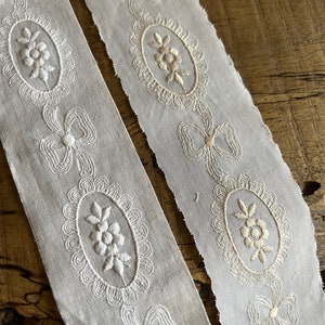 May include: Two white cotton ribbon trims with embroidered floral designs and bows. The trim is delicate and intricate, with a scalloped edge.