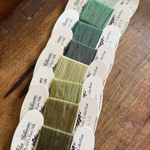 May include: A collection of Elsa Williams 7-Ply 100% Pure Silk thread cards in various shades of green. Each card displays a different color of silk thread, with the card labels visible. The cards are arranged on a wooden surface.