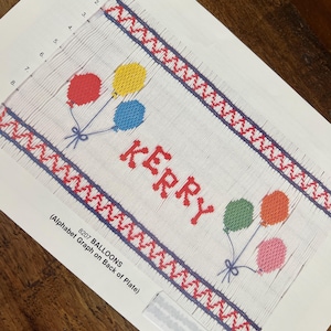 May include: A cross-stitch pattern on paper featuring the name "KERRY" in red letters, with colorful balloons. The design includes red and blue borders with a zig-zag pattern. The text "8207 BALLOONS" is also visible.