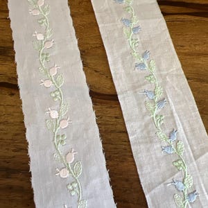 May include: Two strips of white fabric with embroidered floral designs. One strip features pink and green floral patterns, while the other has blue and green floral patterns. The embroidery is delicate and detailed, creating a vintage aesthetic.