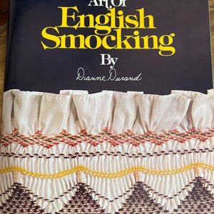 May include: A book cover with the title "The Art of English Smocking" in yellow lettering on a dark blue background. The cover features a close-up of smocked fabric in white, yellow, brown, and red.