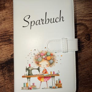 Könnte beinhalten: Weißes Kunstleder-Journal mit dem Wort "Sparbuch" in schwarzer Schrift. Das Cover zeigt eine farbenfrohe Illustration eines Mädchens, das mit einer alten Nähmaschine näht, umgeben von Garnrollen und Knöpfen. Ein weißer Riemen sichert das Journal.