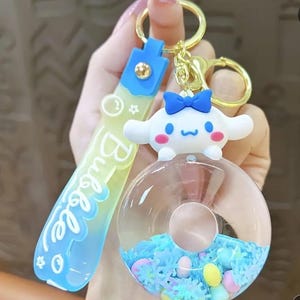 May include: A blue and yellow keychain with the word "Bubble" written on it. The keychain features a clear plastic charm with a donut shape filled with blue and white glitter and colorful sprinkles. A white character with a blue bow is attached to the top of the charm.