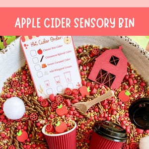 May include: A sensory bin filled with red and brown filler, featuring a red wooden barn, a wooden spoon, two red paper cups with black lids, white pom-poms, and red apple-shaped pieces. A Hot Cider Order card lists items like Classic Hot Cider.