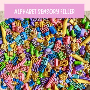 May include: A wooden bowl filled with a colorful sensory filler consisting of various pasta shapes, including alphabet letters, penne, and rotini, mixed with small round beads in shades of blue, green, pink, yellow, purple, and gold.