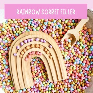 May include: A wooden rainbow-shaped toy with a scoop rests on a bed of colorful, round beads. The text "RAINBOW SORBET FILLER" is displayed in a pink banner at the top. The beads are in various pastel colors.