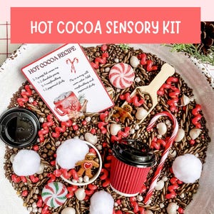 May include: A hot cocoa sensory kit featuring a white bowl filled with brown filler, red and white peppermint candies, white cotton balls, a small wooden scoop, a red paper cup with a black lid, and a card labeled HOT COCOA RECIPE with instructions.