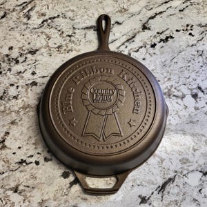 May include: A cast iron skillet with a brown finish. The skillet has a handle and a circular logo that reads "Blue Ribbon Kitchen Country Living Est. 1974".