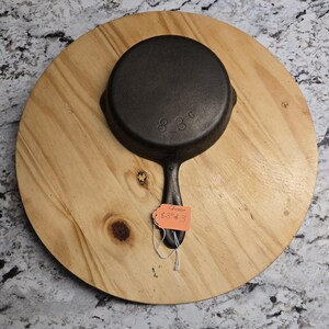 May include: A small, black cast iron skillet with a long handle, resting on a round wooden cutting board. The skillet has the numbers "83 G" embossed on the bottom. A price tag is attached to the handle, with the text "Cheese 8.25 #3".