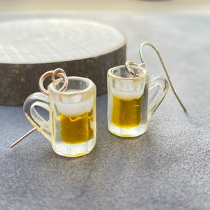 May include: A pair of novelty earrings featuring miniature beer mugs. The clear resin mugs are filled with yellow liquid, topped with white foam, and have silver-colored ear wires. The earrings are designed to resemble miniature beer steins.