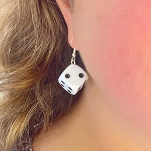 May include: White dice shaped earrings with black dots. The dice have a two on the visible side.