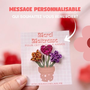 Peut inclure: Une carte carrée avec un motif quadrillé et une illustration de plante en pot avec trois formes pailletées : une flamme orange, un cœur rose et une étoile violette. Le texte indique : Merci Maîtresse POUR CETTE BELLE ANNÉE et ATELIERROCHELAIS.FR.