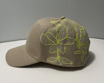 Op de afbeelding: Een beige baseballpet met een neon-groen geborduurd bloemenontwerp aan de zijkant.
