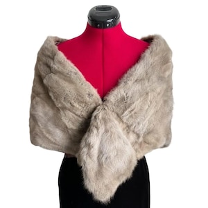 May include: A light brown fur shawl draped over a red mannequin. The shawl is made of soft, textured fur and is designed to be worn over the shoulders. The garment is displayed against a white background.