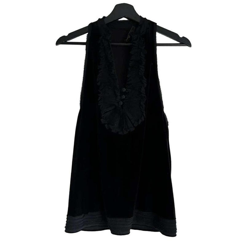 Puede incluir: Top negro sin mangas con cuello en V y ribete decorativo de volantes. La parte superior tiene tres botones peque&ntilde;os y un dobladillo gris en contraste. La prenda est&aacute; colgada de una percha negra.