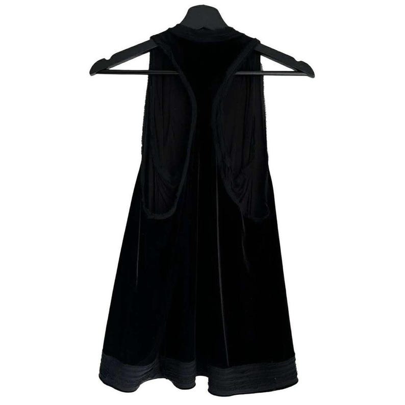 Puede incluir: Un chaleco de terciopelo negro cuelga de una percha negra. El chaleco tiene un cuello alto y un dise&ntilde;o frontal abierto. La tela parece suave y cae con elegancia. El dobladillo inferior est&aacute; adornado con un material m&aacute;s oscuro, similar al sat&eacute;n.